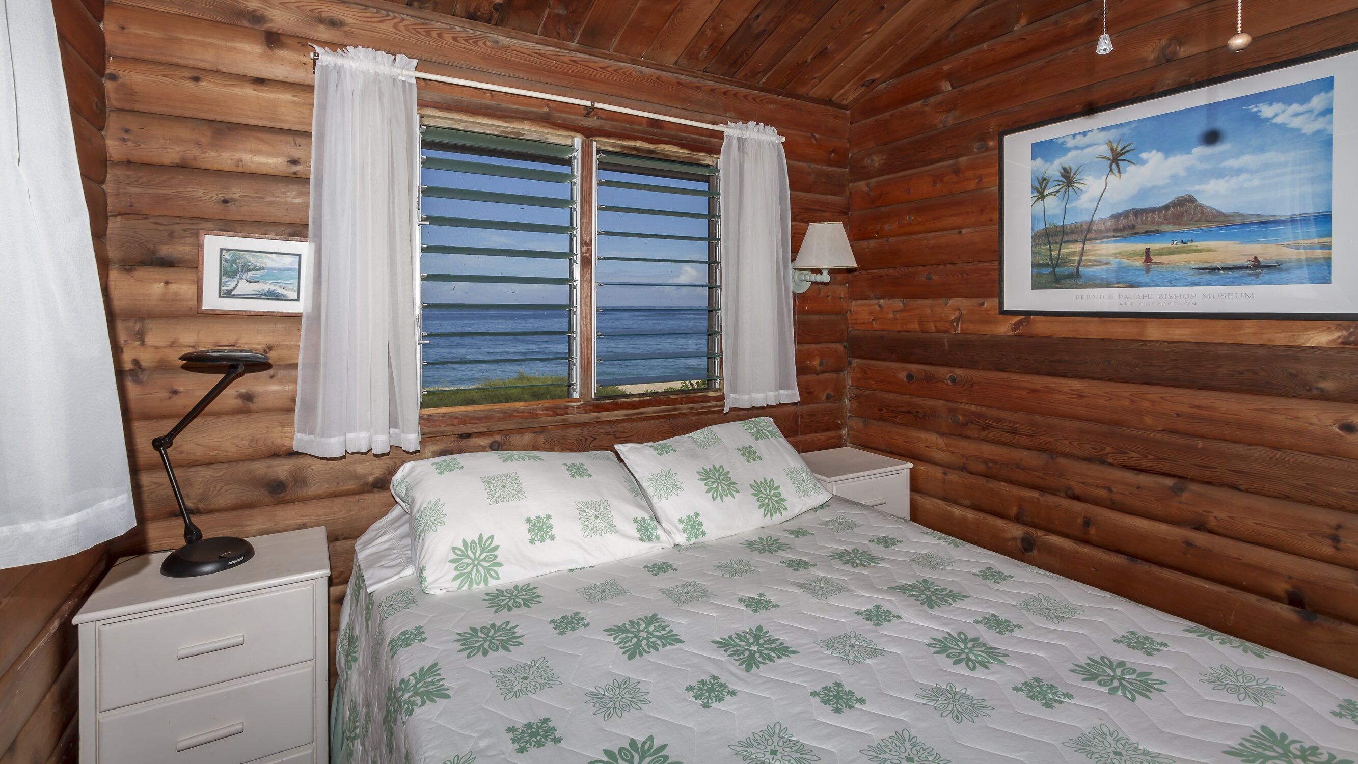 A wooden cabin room with a bed, a nightstand, a lamp, and a window with curtains.