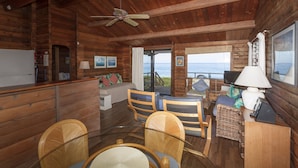 A cabin interior with wooden walls, a ceiling fan, a dining area with wicker chairs, a sofa, and a television. - Beachfront Cottage at Log Cabins (Haleiwa)