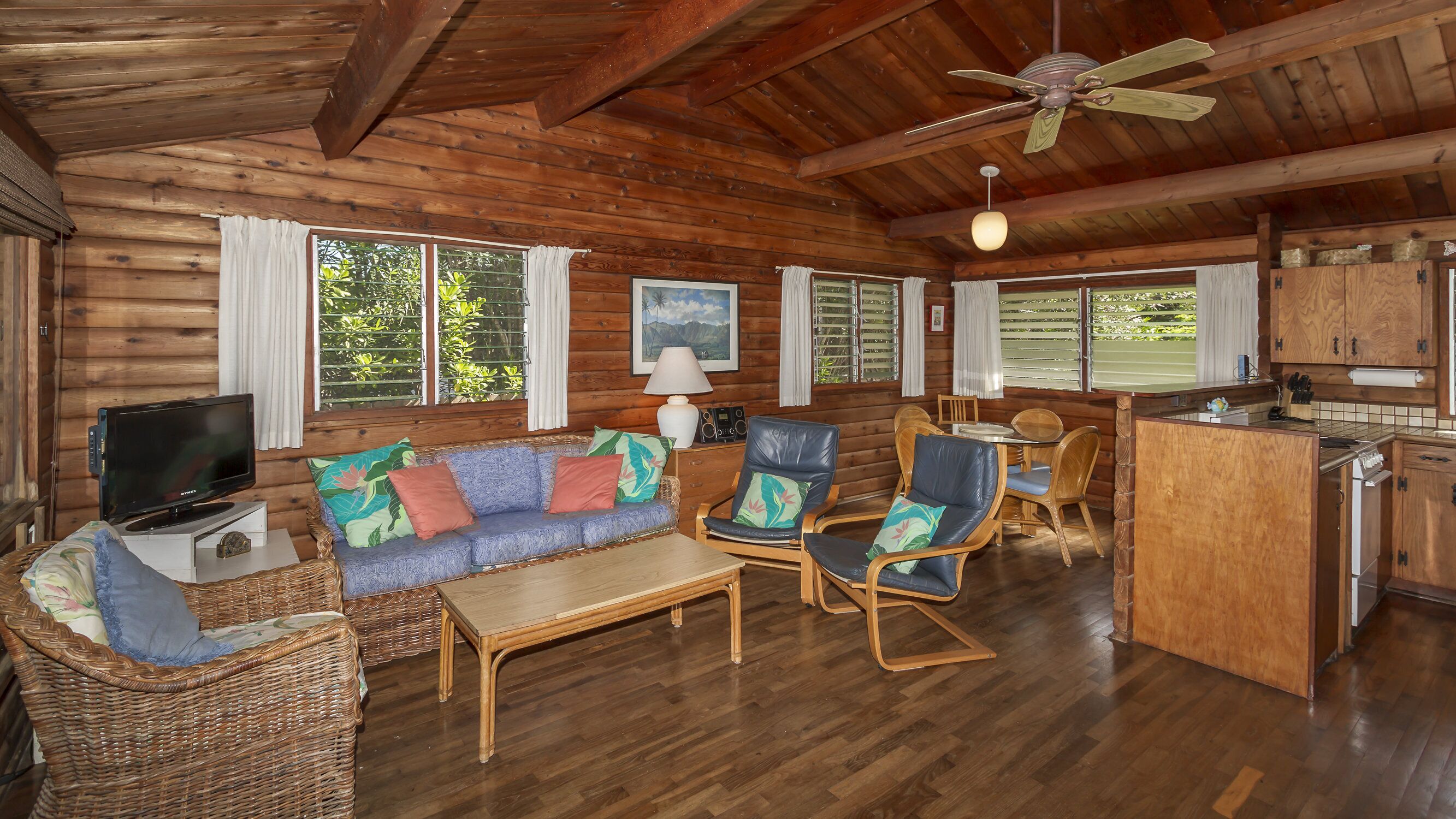 A wooden cabin interior with a sofa, chairs, a television, a dining table, and a kitchen area.