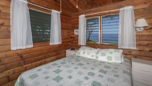 A wooden cabin bedroom with a bed, two windows with curtains, a nightstand, and a lamp. - Beachfront Cottage at Log Cabins (Haleiwa)