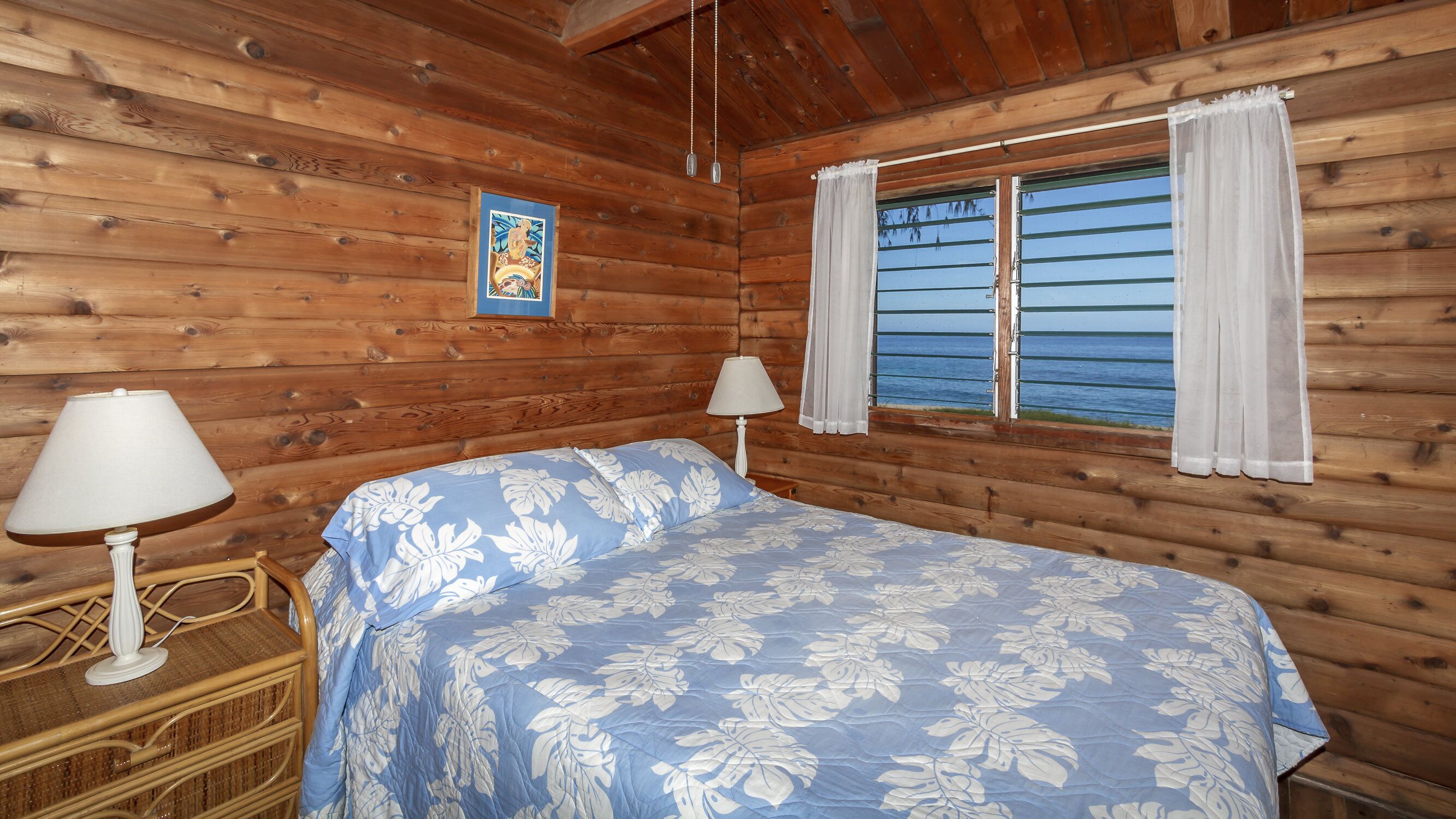 Ein Zimmer in einer Holzhütte mit einem Bett, zwei Nachttischlampen, einem Fenster mit Vorhängen und einem gerahmten Bild an der Wand.