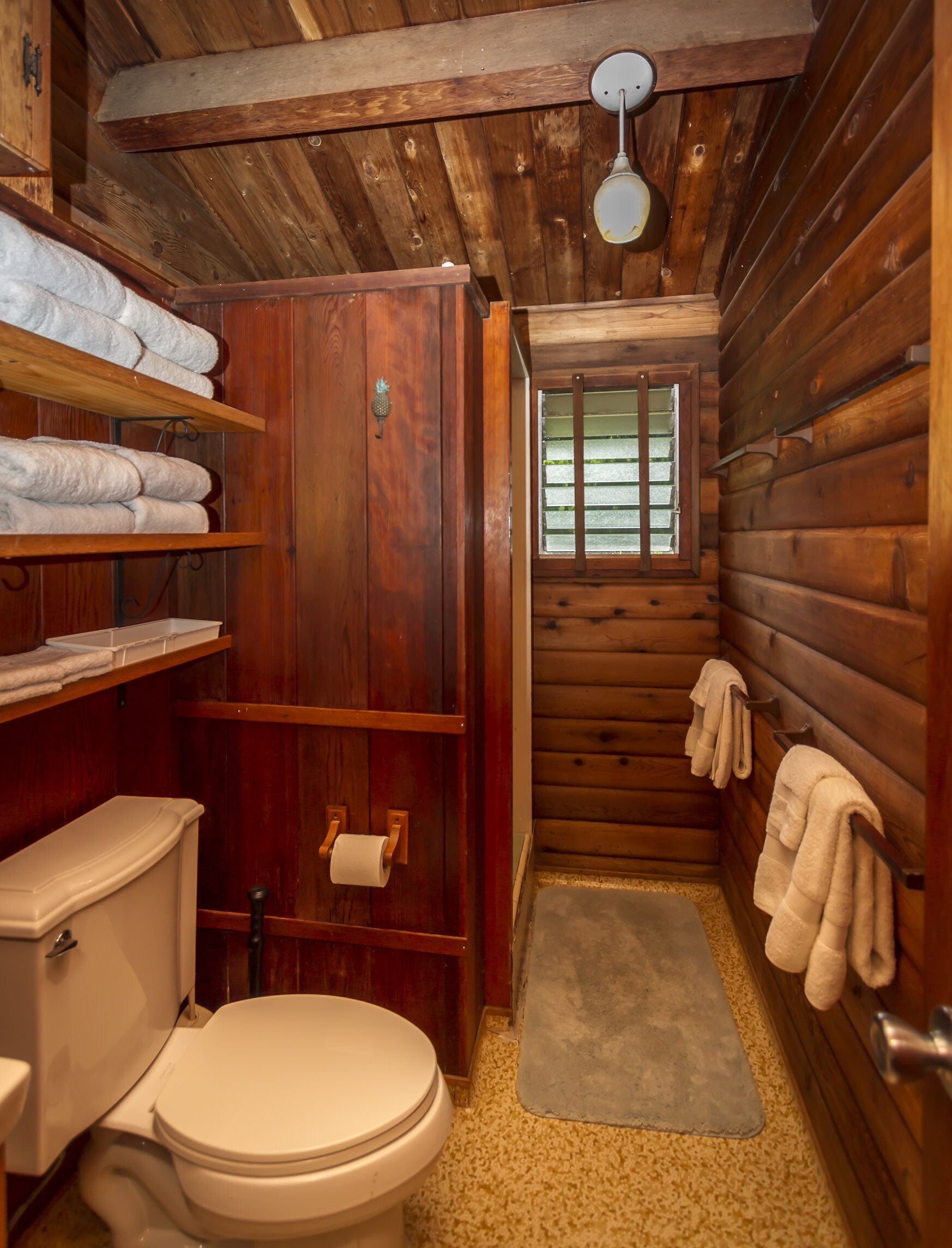 A compact wooden cabin bathroom with a toilet, shelves with towels, a window, and a wooden ceiling.