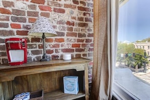 A room with a brick wall, a wooden table with a coffee maker, a lamp, and a window with a view of buildings and a street.
