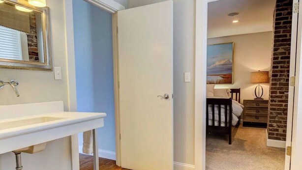 A bathroom with a sink, mirror, and door leading to a bedroom with a bed, dresser, and a painting on the wall. - The Soho Suite - Where Luxury & Location Meet (Charleston)