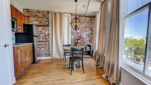 A kitchen with a brick wall, wooden cabinets, a refrigerator, a dining table with chairs, and a window with a view. - The Soho Suite - Where Luxury & Location Meet (Charleston)