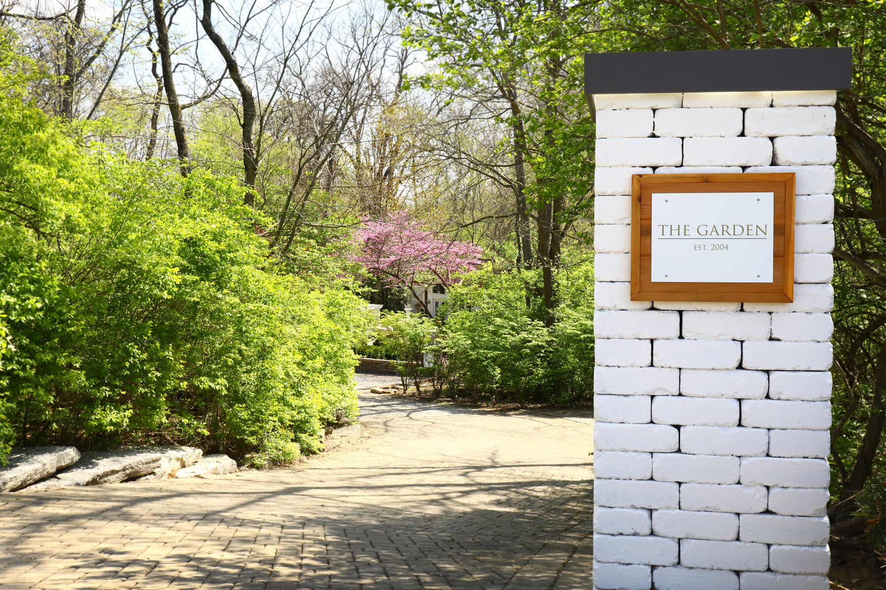 A brick pathway leading to a white wall with a sign that reads "THE GARDEN EST. 2004".
