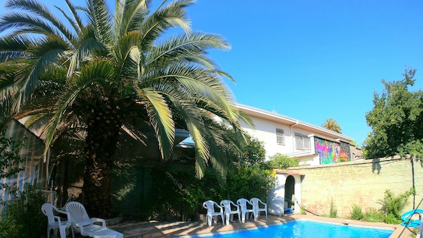 A swimming pool with white lounge chairs, a palm tree, and a house with a colorful mural. - Hotel Genova (Los Andes)