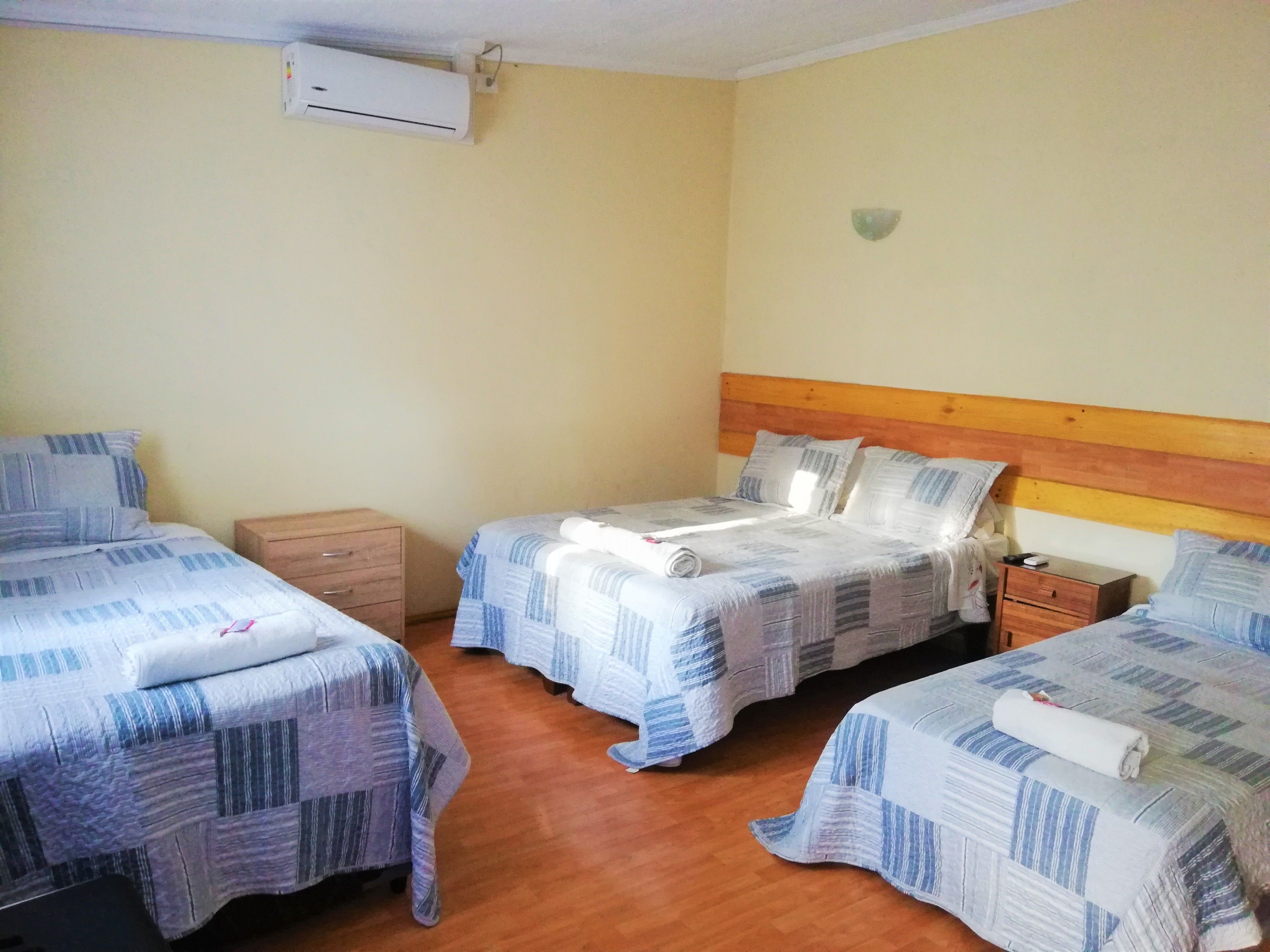 Una habitación con tres camas, cada una con cabecero de madera, una mesita de noche de madera y un aire acondicionado instalado en la pared.