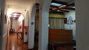 A hallway with wooden flooring, white walls, and arched doorways. There is a fire extinguisher on the left, a clock on the wall, and a radiator on the right. - Hotel Genova (Los Andes)