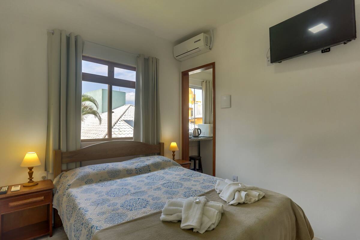 A hotel room with a bed, a TV mounted on the wall, an air conditioning unit, a window with curtains, and a view of buildings outside.
