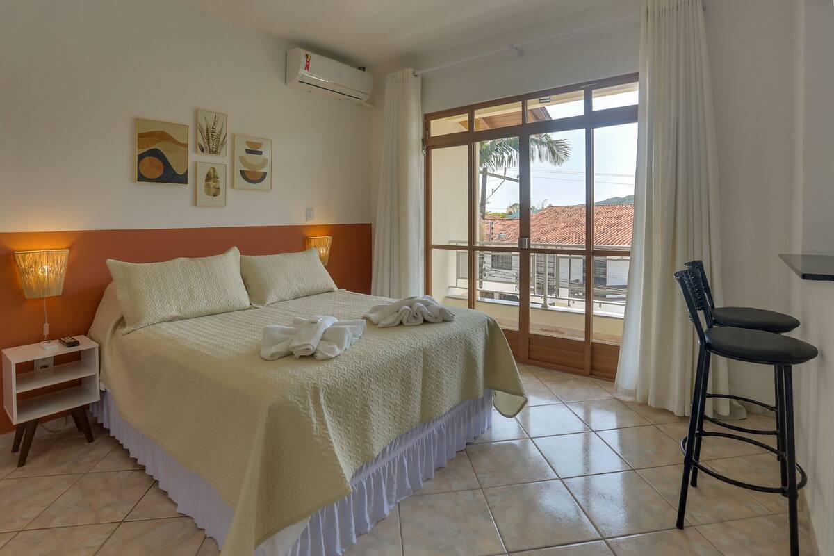 A hotel room with a bed, a nightstand, a chair, and a large window with a view of buildings and palm trees.