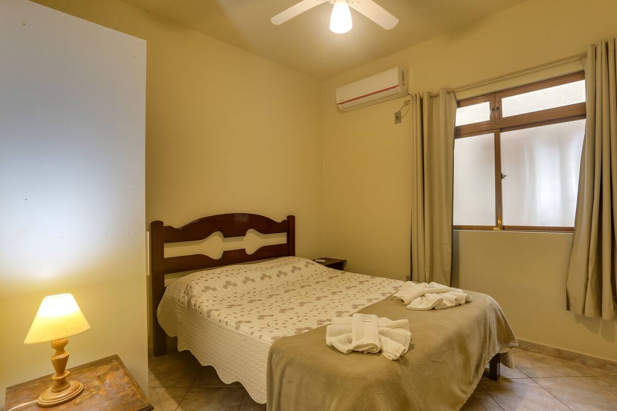 A bedroom with a bed, a nightstand, a lamp, an air conditioning unit, and a window with curtains.