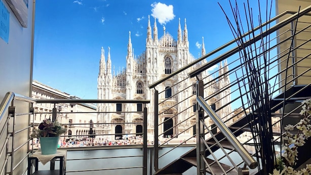 Vista de una gran catedral a través de un amplio ventanal desde un balcón con barandilla de metal y una pequeña mesa con planta en maceta.
