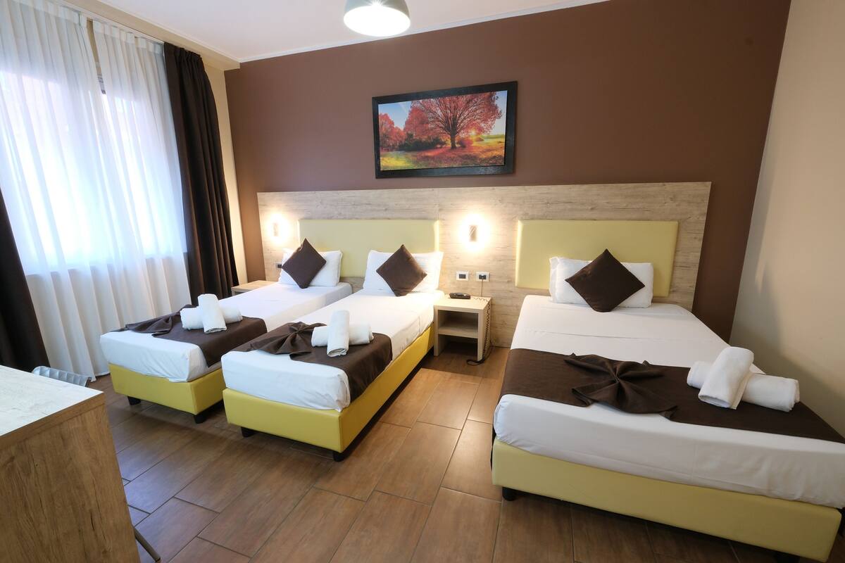 A hotel room with three beds, a wooden headboard, a painting of a tree, and a nightstand with a lamp.