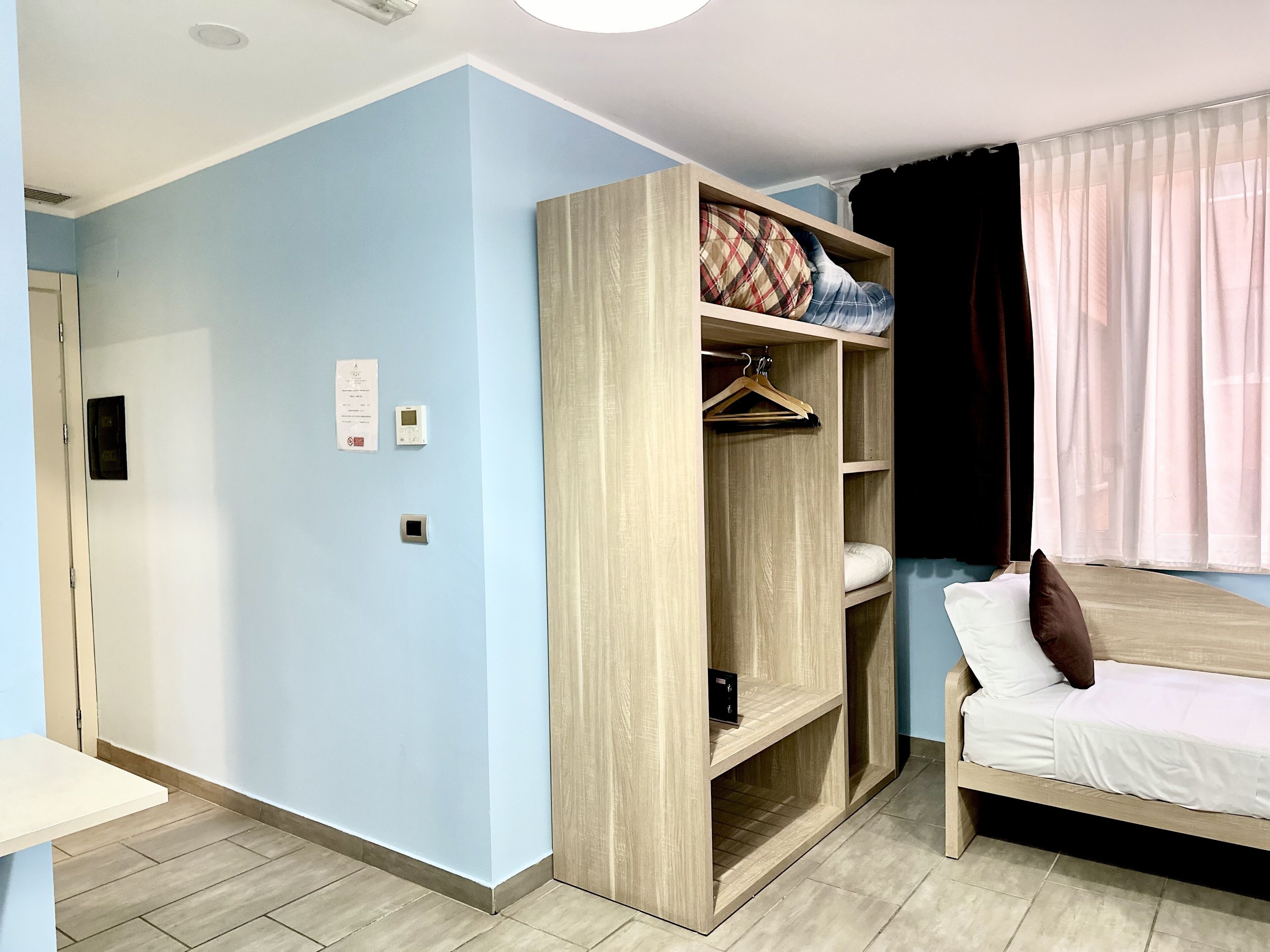 A bedroom with a light blue wall, a wooden wardrobe, a bed with white bedding, and a window with sheer curtains.