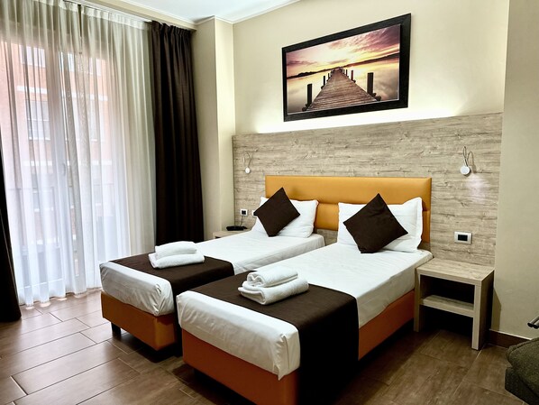 Habitación de hotel con dos camas, cabecero de madera, un cuadro de un atardecer y vistas a un edificio a través de cortinas translúcidas.