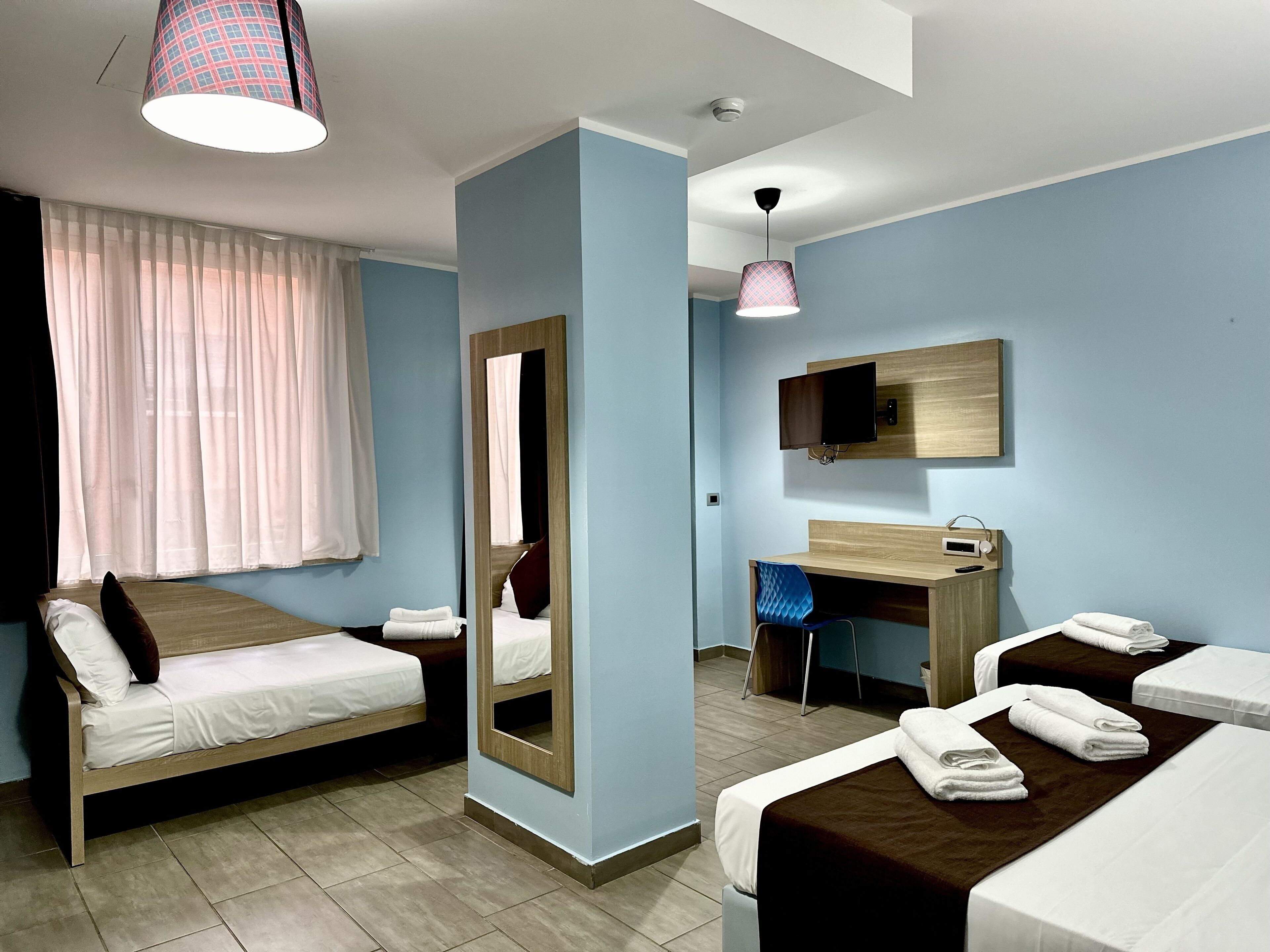 A hotel room with two single beds, a desk with a chair, a mirror, a TV, and a ceiling light.