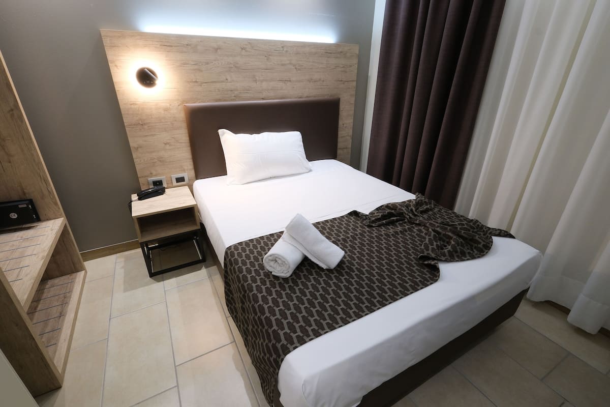 A hotel room with a bed, a bedside table, a nightstand, a wall-mounted light, and a window with curtains.