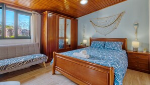 Un dormitorio con techo de madera, una cama con colcha azul estampada, una banca de madera y una ventana con cortinas.