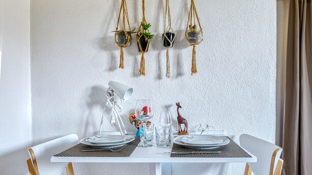 Un comedor con una mesa blanca, dos sillas de madera, una planta decorativa colgante y una pequeña estatua sobre la mesa.