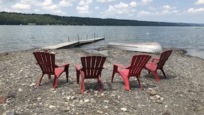 Een landschap bij een meer met rode Adirondack-stoelen op een rotsachtige oever, een houten steiger die het water in loopt en een boot op de steiger.
