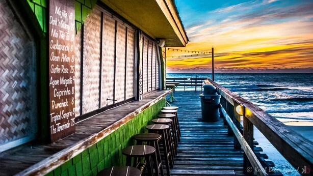 Een restaurant aan zee met een houten terras en uitzicht op de oceaan tijdens de zonsondergang.