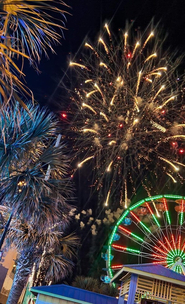 A night scene with fireworks, a Ferris wheel, and palm trees. - ~ Off-Season Rental - SeaMist Seas - Pool, Steps to Oceanfront Tiki Bar (Carolina Beach)