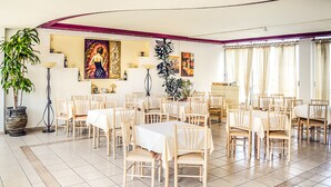 A spacious dining area with multiple tables and chairs, a large potted plant, and artwork on the walls. - Alexandros Hotel (Agios Nikolaos)