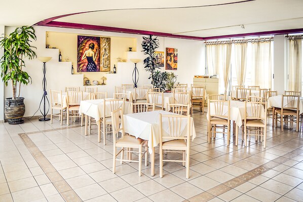 A spacious dining area with multiple tables and chairs, a large potted plant, and artwork on the walls. - Alexandros Hotel (Agios Nikolaos)