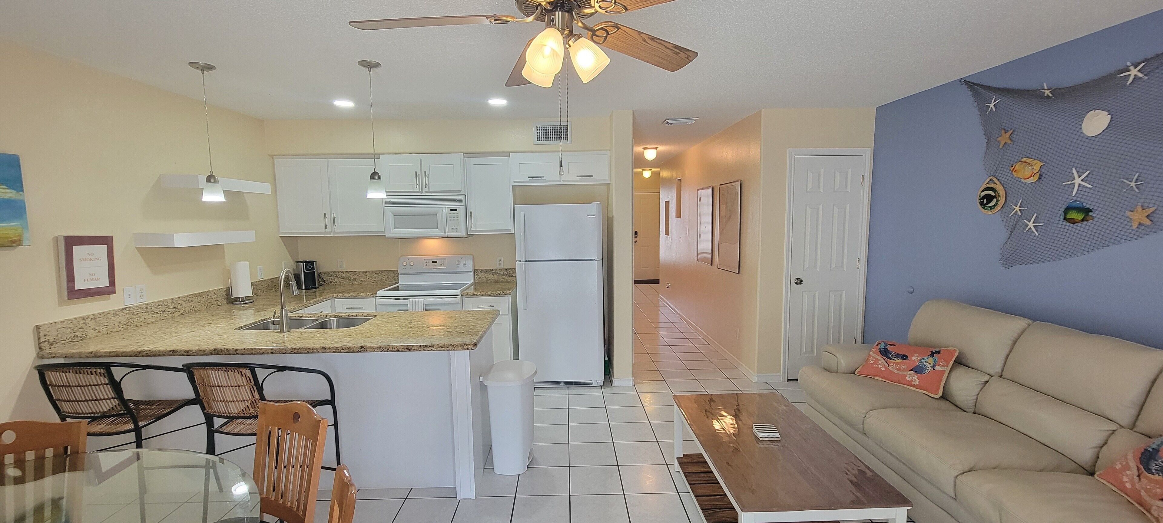 A compact kitchen with white appliances, a dining area with a glass table and wicker chairs, and a living room with a beige sofa and a decorative wall.