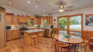 A kitchen with wooden cabinets, a central island with stools, a ceiling fan, and a large window with a view of greenery. - Lakefront Country Haven near Ocala & Forest -With Canoe, Lifejackets and Grill! (Ft McCoy)