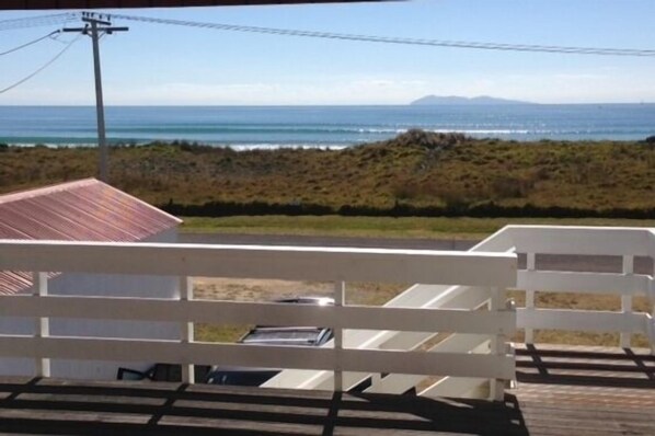 A view from a covered deck overlooking the ocean, with a white fence in the foreground and a building with a red roof in the background.