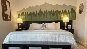 A neatly made bed with a mountain-themed wall art, two bedside lamps, and three storage bins under the bed. - Sherpa Studio (Whitefish)