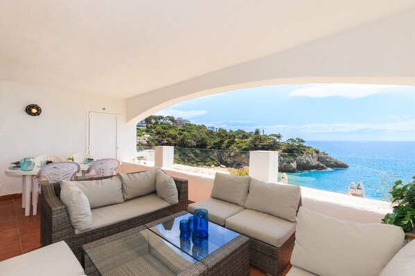Een balkon met rieten meubilair, een glazen tafel en uitzicht op zee en groen.