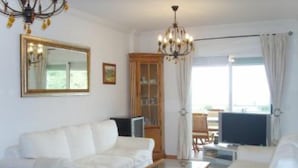 Ein Wohnzimmer mit einem weißen Sofa, einem Holztisch, einer Kristallleuchtre, einem großen Spiegel und Blick ins Freie durch ein Fenster mit Vorhängen.