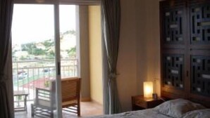 Ein Schlafzimmer mit Bett, Deckenventilator, Nachttisch mit Lampe und Blick durch eine Glasschiebetür auf einen Außenbereich mit Stuhl und Tisch.