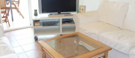 Una sala de estar con una mesa de centro de vidrio, un sofá blanco, un televisor sobre un mueble bajo y un comedor con sillas.