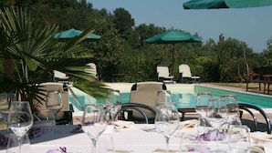 A table set for a meal with wine glasses, plates, and cutlery, overlooking a swimming pool with lounge chairs and umbrellas.