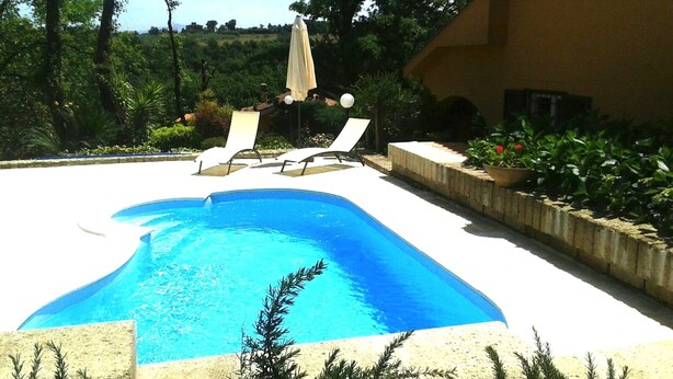 A swimming pool with lounge chairs and an umbrella, surrounded by greenery and a building. - Charming Villa with pool near Rome  (Sacrofano)