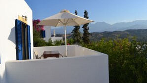 A white building with blue shutters, a table, and an umbrella on a balcony overlooking a mountainous landscape. - Complex of apartments & studio, full-equipped, in a large, beautiful garden (Kamilari)