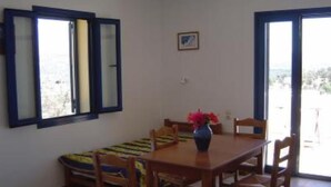 A room with a wooden table and chairs, a small sofa, a vase with flowers, and a window with blue frames. - Complex of apartments & studio, full-equipped, in a large, beautiful garden (Kamilari)