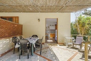 A covered patio area with a dining table and chairs, a kitchen visible through an open doorway, and a wooden fence on the left. - Beautiful location overlooking the sea. (Erice)