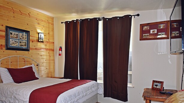 A bedroom with a wooden wall, a bed with a red blanket, a small table with a picture frame, and a window with curtains. - Bear Trap Apartments - Full Kitchen (Gardiner)