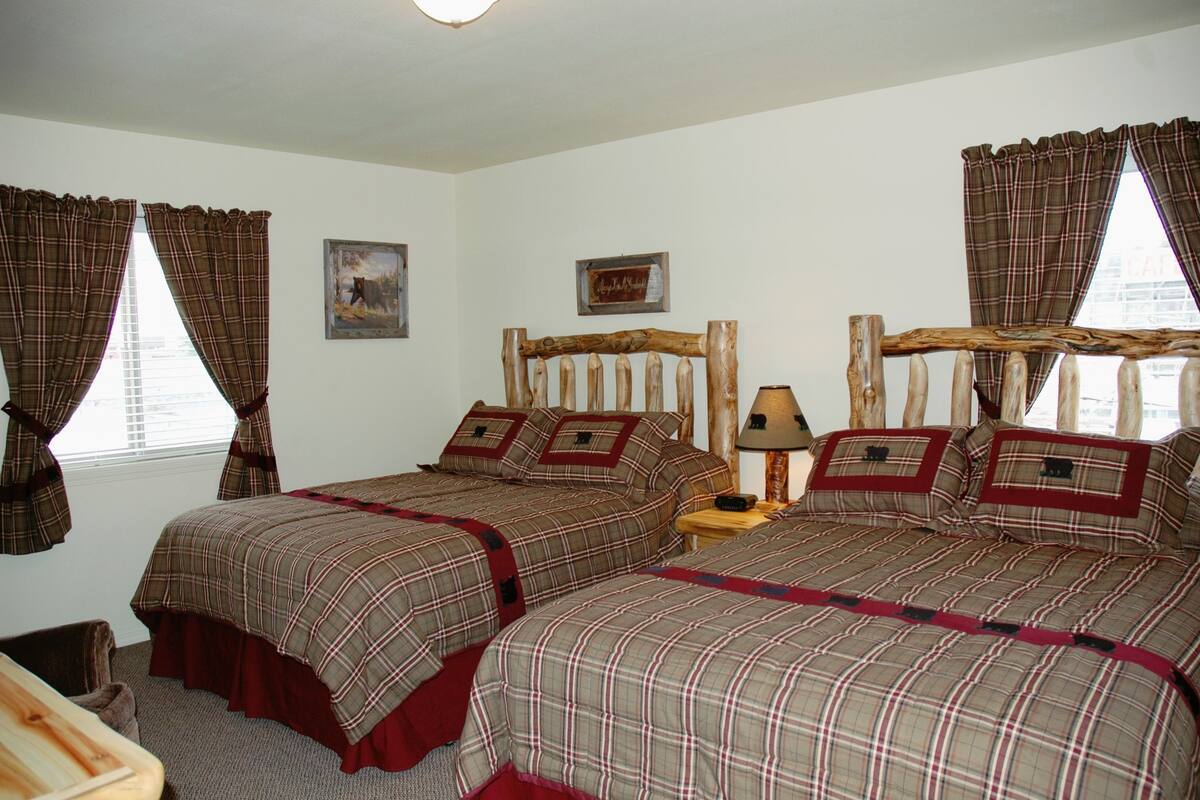 A bedroom with two beds, plaid bedding, a wooden headboard, a nightstand, and a lamp.