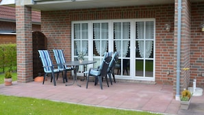 Een terras met een bakstenen muur, een overdekt zitgedeelte met gestreepte stoelen en een tafel, en een groot raam met gordijnen.