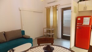 Un espace de vie compact avec un canapé turquoise, une petite table ronde recouverte d’une nappe en dentelle blanche, un réfrigérateur rouge et un banc en bois.