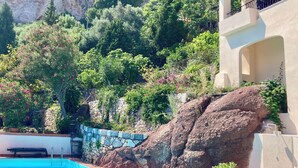 Un área de piscina con un muro de piedra y una gran roca, rodeada de vegetación exuberante y un acantilado.
