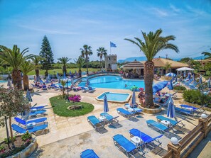 Un complexe hôtelier doté d’une grande piscine entourée de chaises longues et de parasols, d’une structure ressemblant à un château et de palmiers.