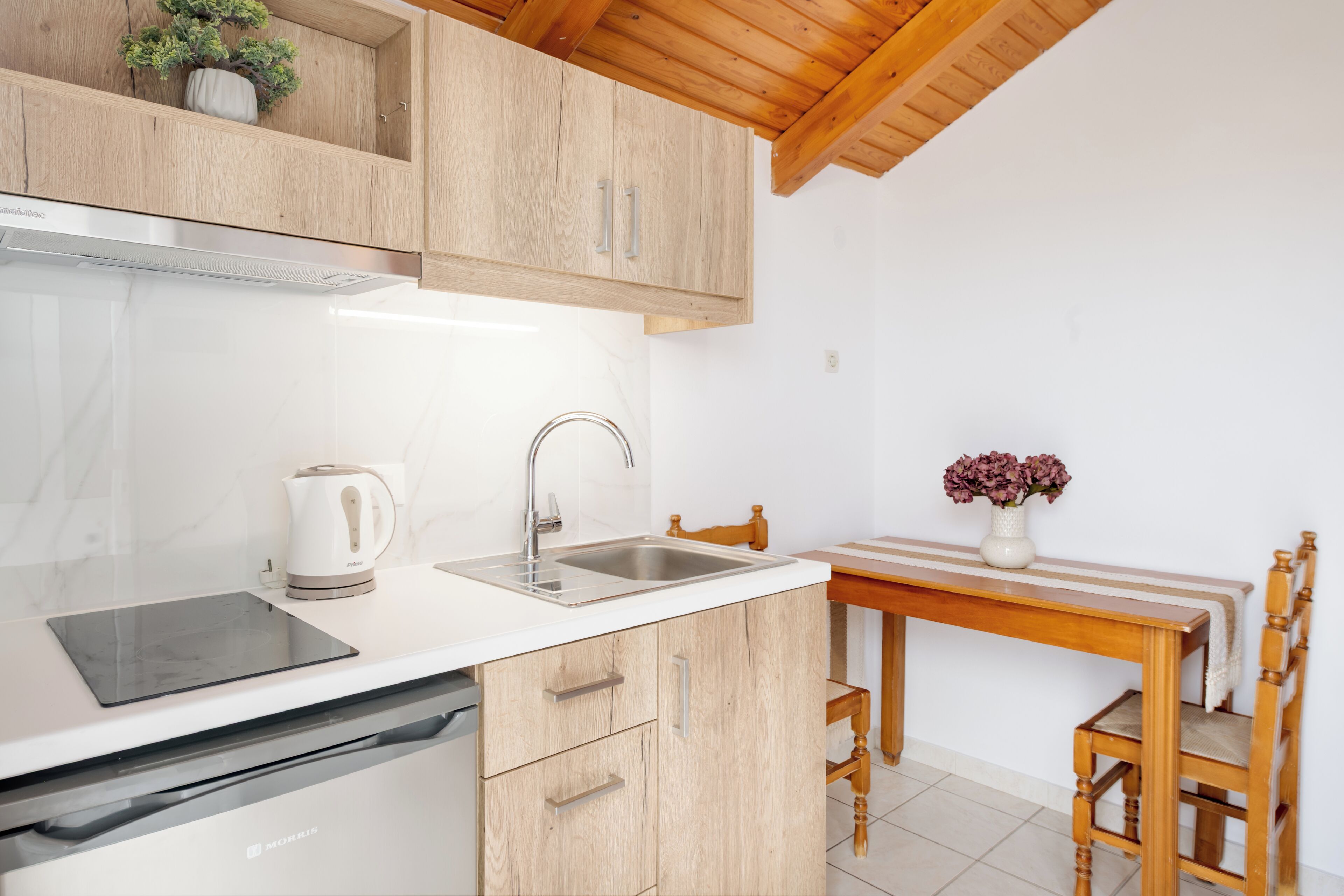 Een keuken met houten kastjes, een roestvrijstalen spoelbak, een waterkoker en een eettafel met stoelen.