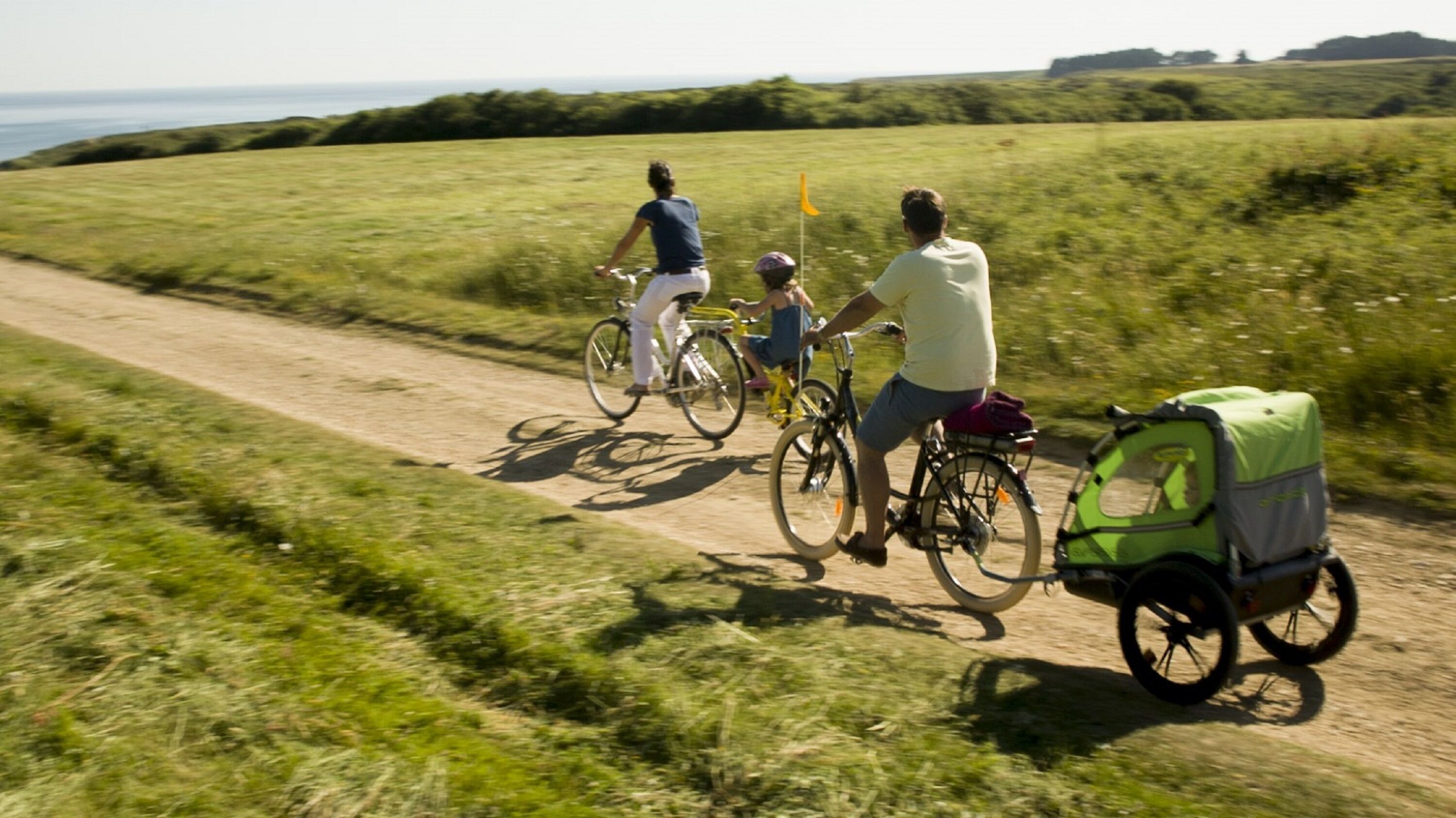 A dirt path with cyclists and a child, surrounded by green fields and distant hills.